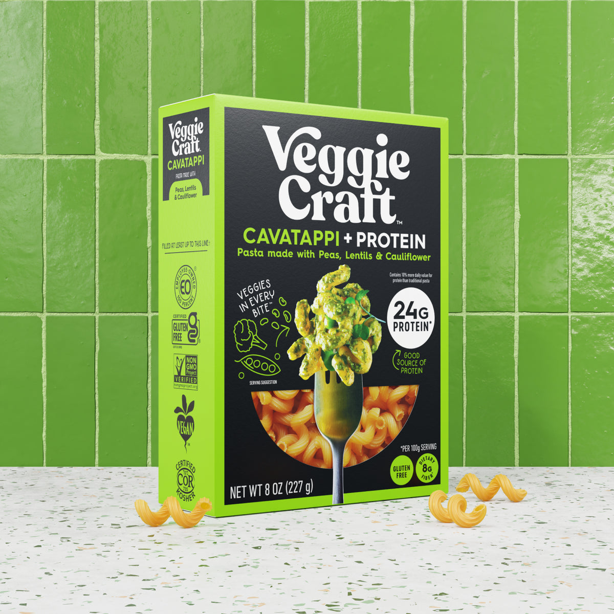 Veggie Craft Cavatappi + Protein pasta box in bright green and black packaging, shown standing on a speckled white surface with loose yellow-orange pasta pieces in front, against a green tiled background.