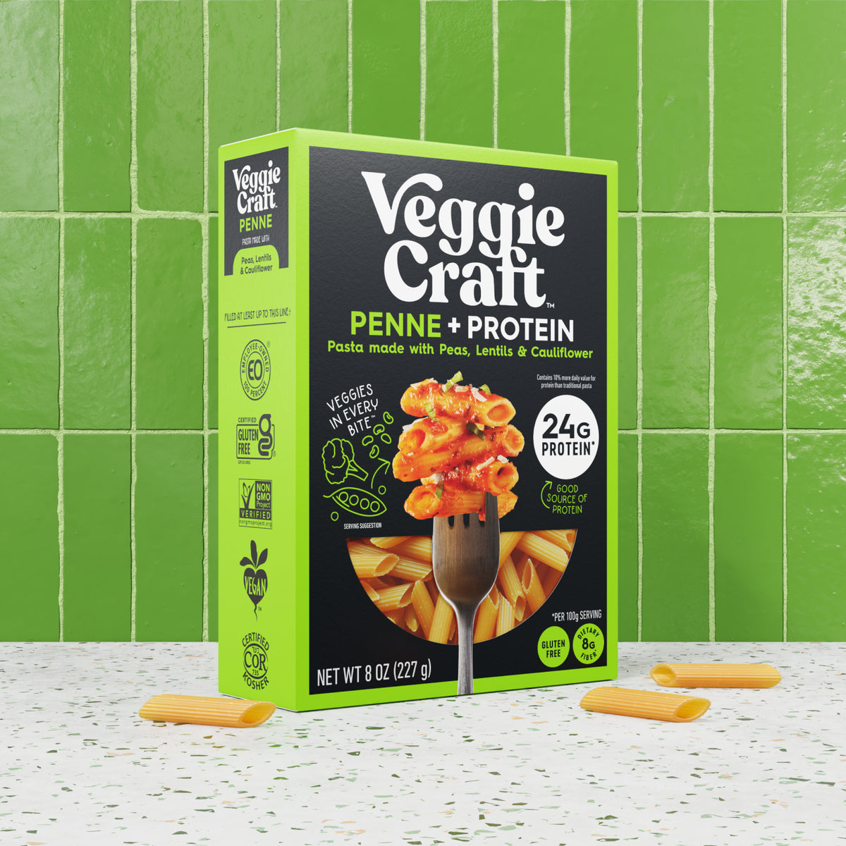 A box of Veggie Craft Penne + Protein pasta stands upright on a speckled white surface with green ceramic tiles behind it. The packaging is bright lime green and black, with a photo of pasta on a fork on the front.