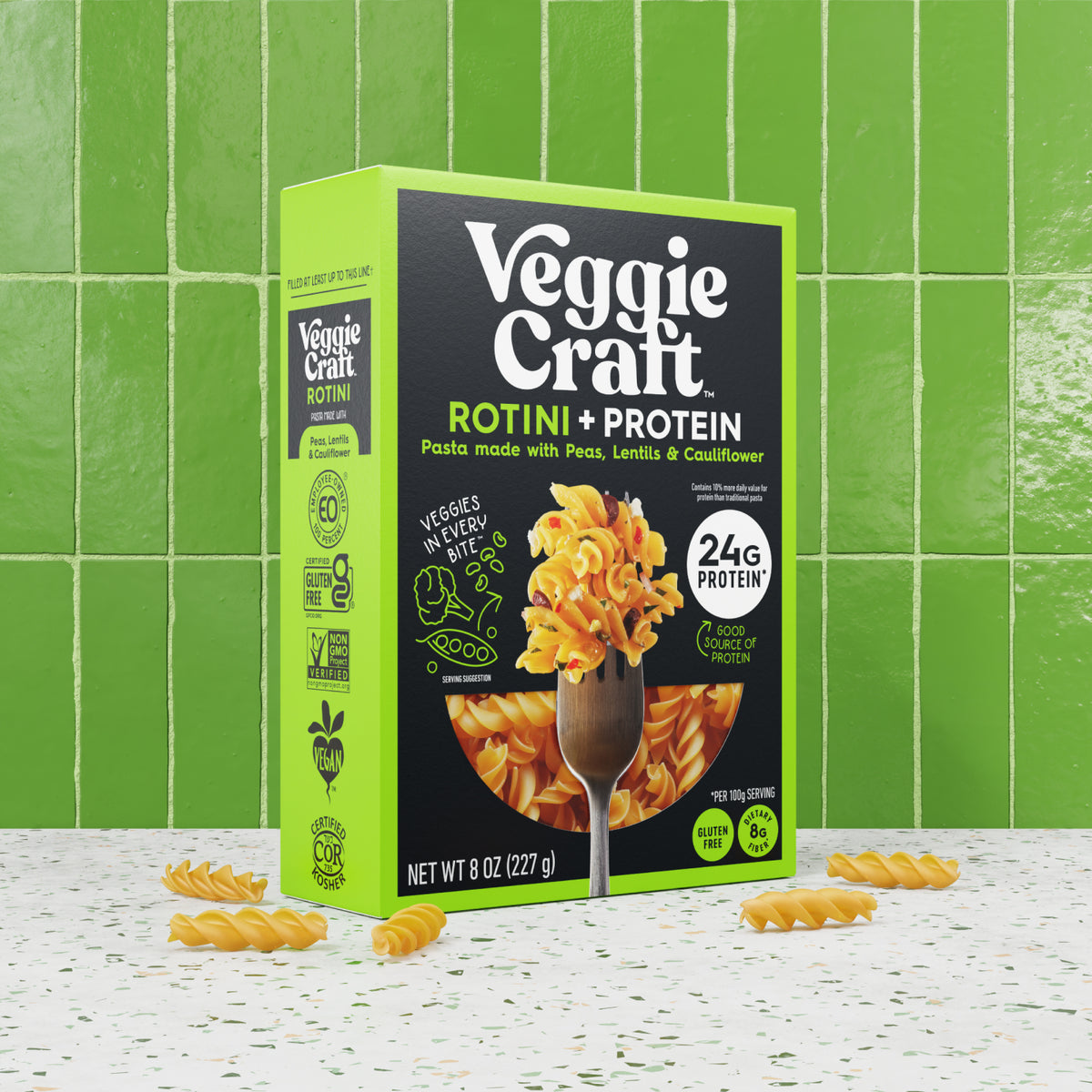 A box of Veggie Craft Rotini + Protein pasta stands on a speckled white surface with loose rotini pieces scattered around it, set against a bright green tiled background.