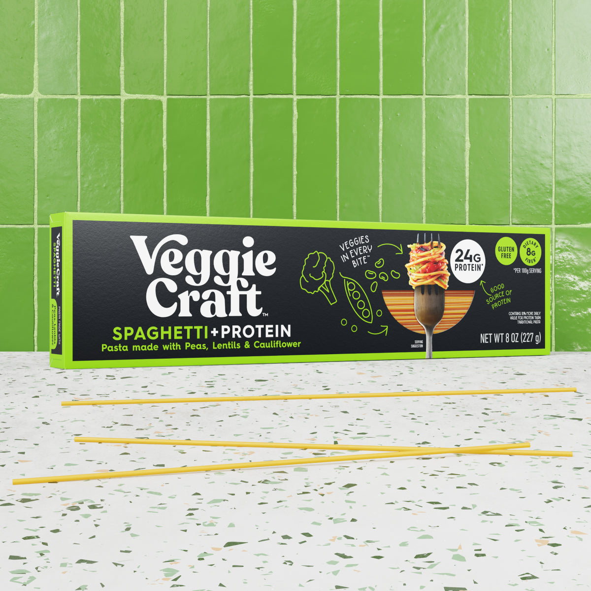A box of Veggie Craft Spaghetti + Protein pasta sits on a speckled white countertop with several yellow spaghetti strands laid out in front. The background features glossy green subway tiles.