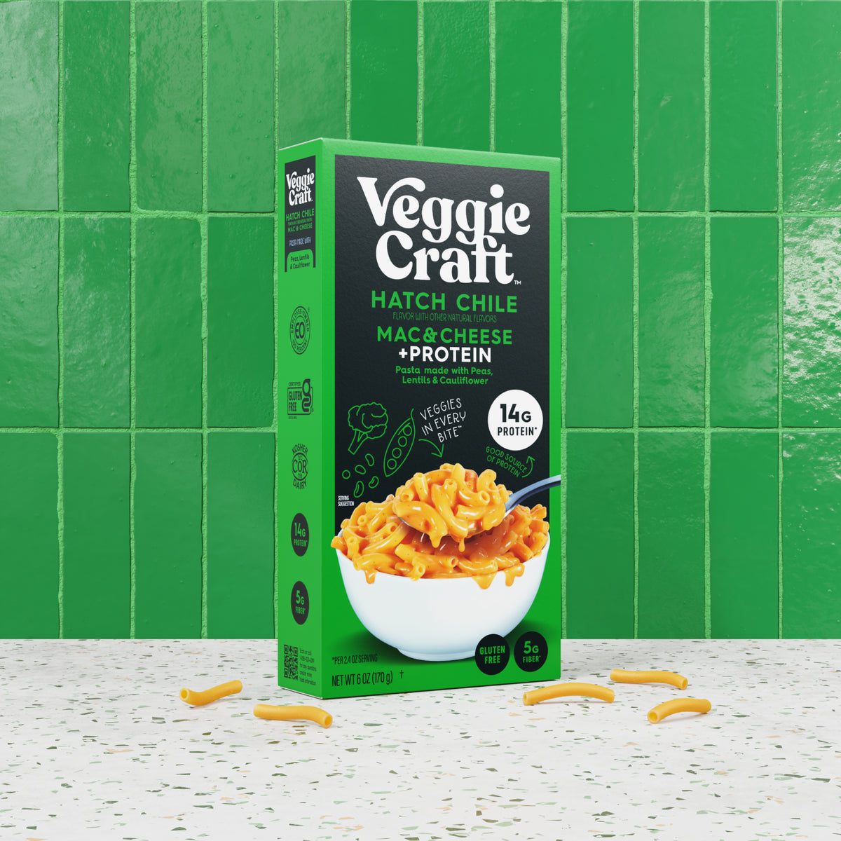 A box of Veggie Craft Hatch Chile Mac & Cheese + Protein stands upright on a speckled white surface against a bright green tiled wall. The package is green and black with white text and an image of a bowl of cheesy macaroni with orange-red chile sauce.