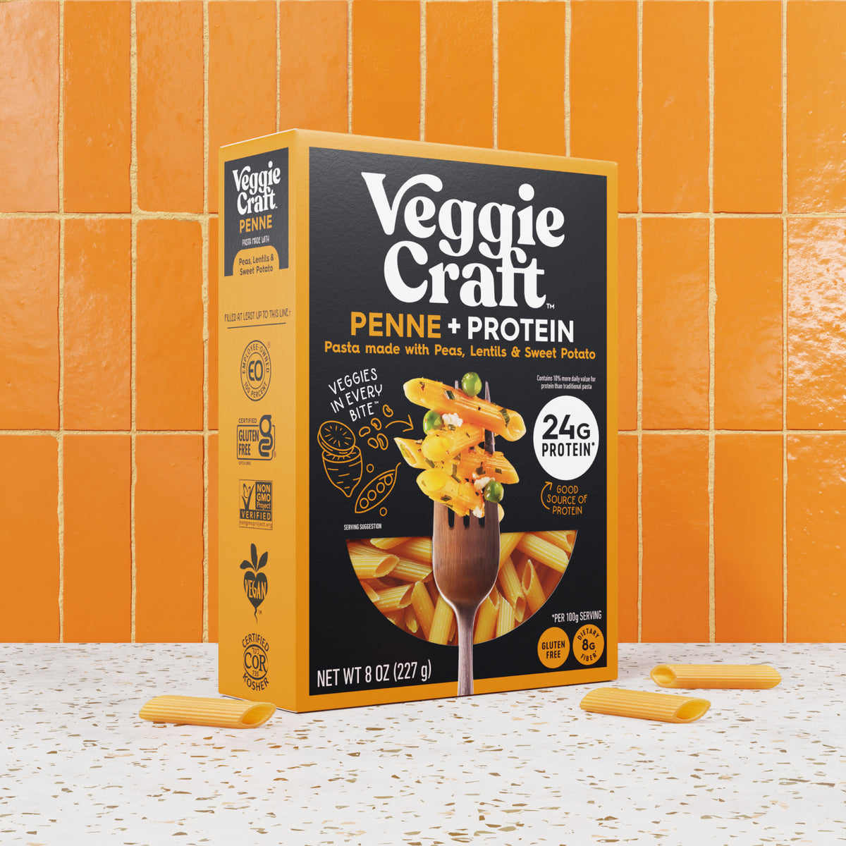 A box of Veggie Craft Penne + Protein pasta stands upright on a speckled white surface with orange ceramic tiles behind it. The package is black and yellow with a photo of pasta on a fork and text including 'Penne + Protein' and '24g protein.
