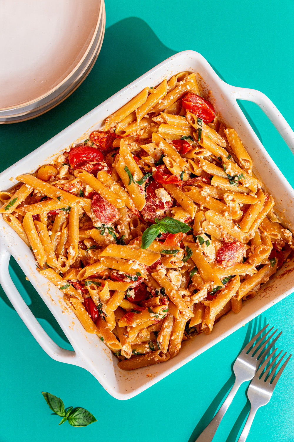 Penne pasta dish with tomatoes and herbs in a white dish on a teal background