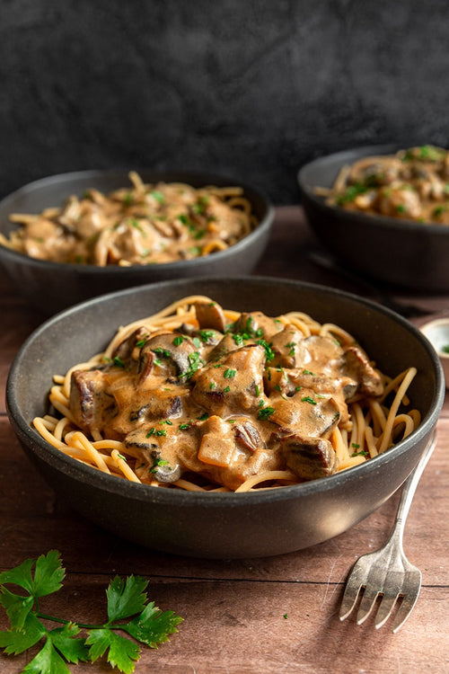 Bowl of creamy mushroom pasta with two additional bowls in the background on a wooden surface.