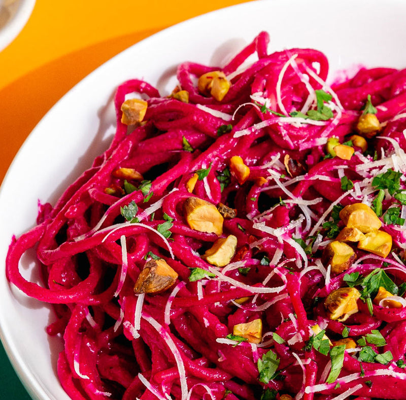 A close-up shot of a vibrant magenta-pink pasta dish, likely beet spaghetti, served in a white bowl. The pasta is garnished with finely chopped nuts, shredded white cheese, and fresh green parsley, set against a bright orange and dark green background.