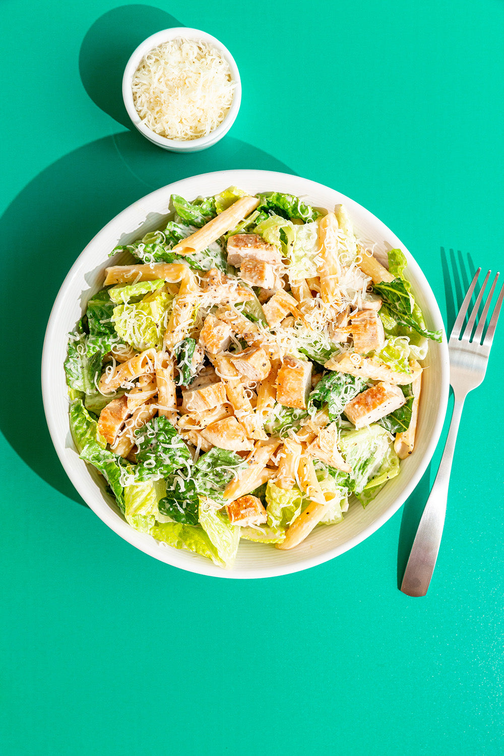 Caesar salad with pasta, croutons, and parmesan cheese on a white plate with a fork on a teal background.