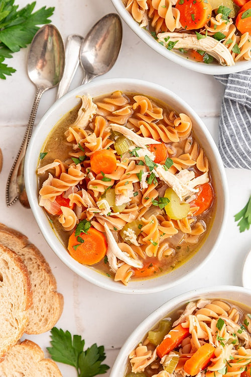 Bowl of chicken noodle soup with carrots, celery, and herbs, served with bread and spoons on a white table.