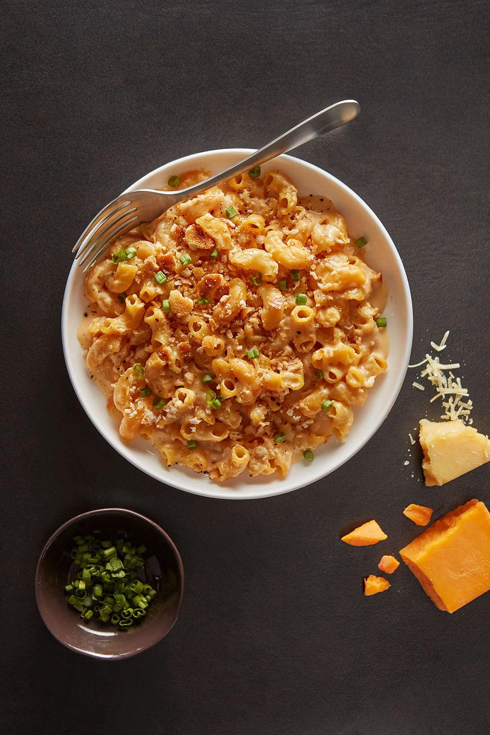 Macaroni and cheese dish with a fork on a dark surface, accompanied by cheese and green herbs.