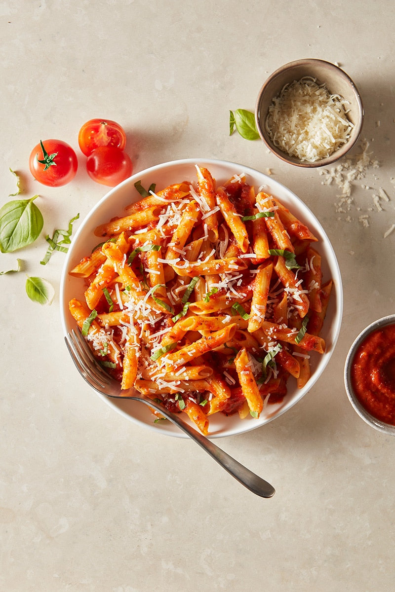 Bowl of penne pasta with tomato sauce, garnished with basil and Parmesan cheese, on a light background.