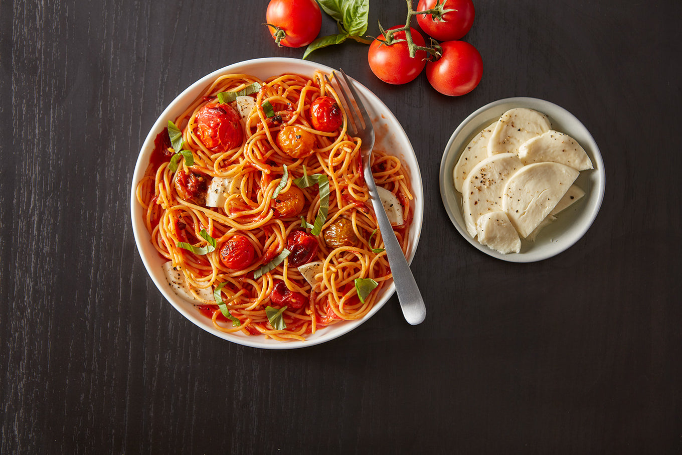 Spaghetti dish with tomatoes and basil in a bowl on a dark surface, accompanied by a small bowl of bread.