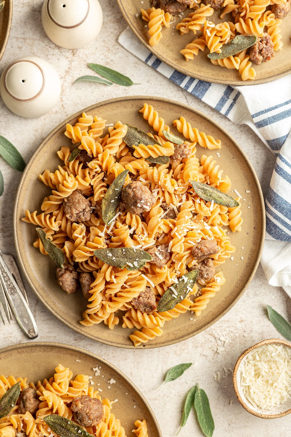 Pasta dish with sausage and sage on a beige plate, served on a light surface with a striped cloth.