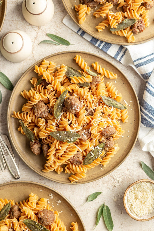 Pasta dish with sausage and sage on a beige plate, served on a light surface with a striped cloth.