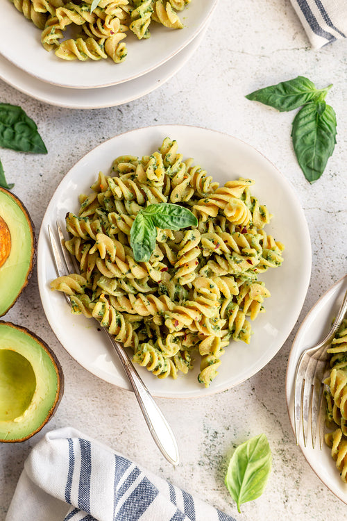 Pasta dish with green sauce on a white plate, surrounded by basil leaves and avocado halves.