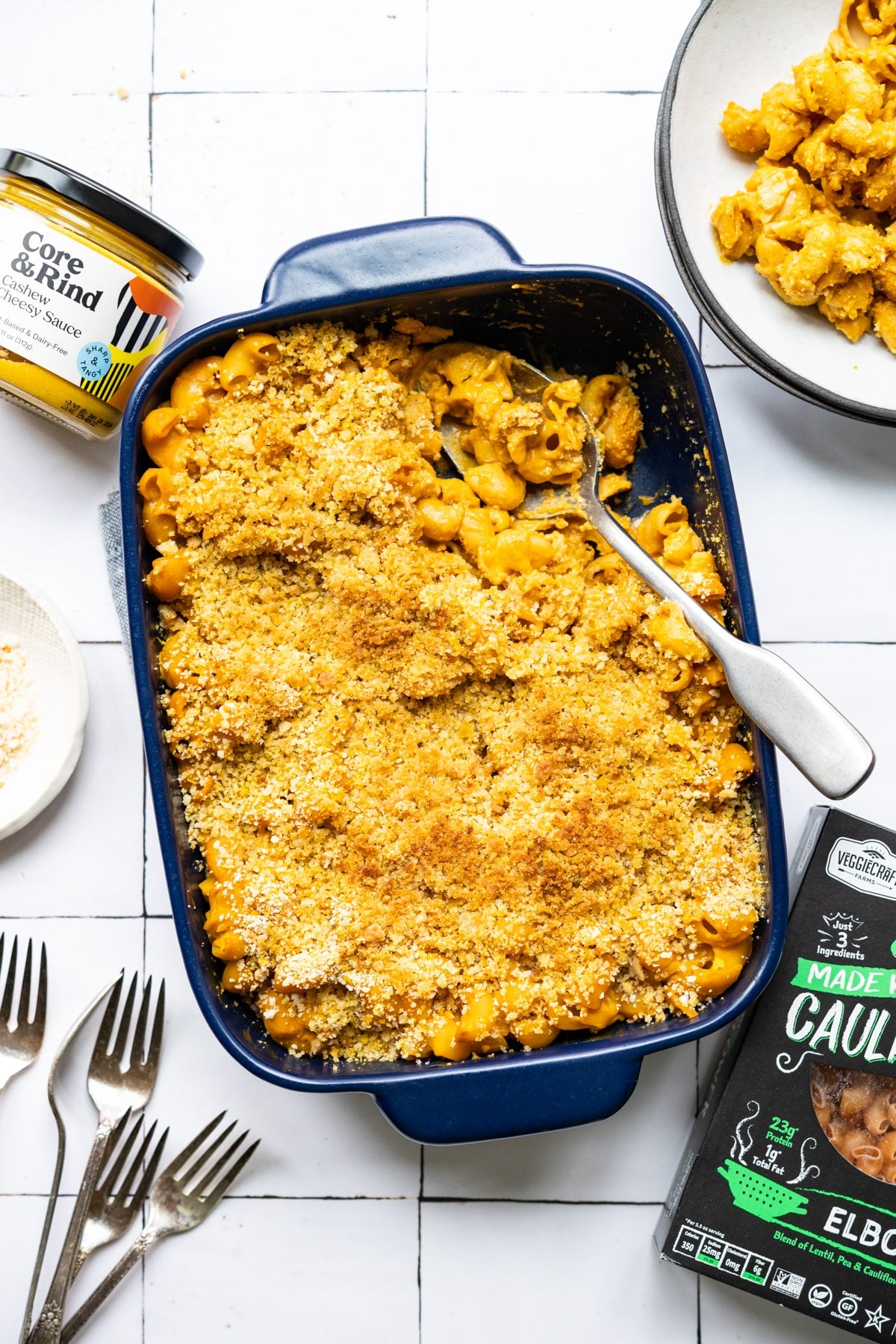 Baked mac and cheese in a blue dish with a can of corn and a package of cauliflower on a white tiled background.