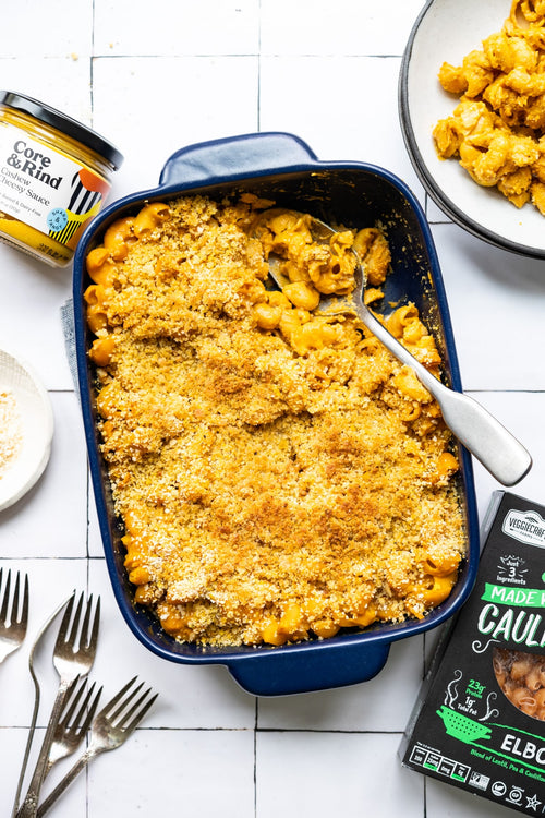 Baked mac and cheese in a blue dish with a can of corn and a package of cauliflower on a white tiled background.