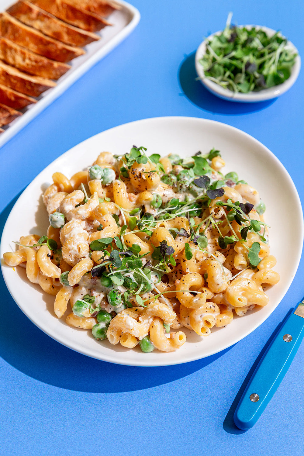 Plate of pasta with green garnish on a blue table with bread and a bowl of greens.