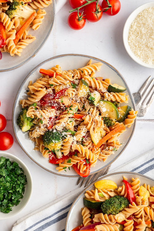 Plates of pasta with vegetables on a white table with tomatoes and Parmesan cheese.