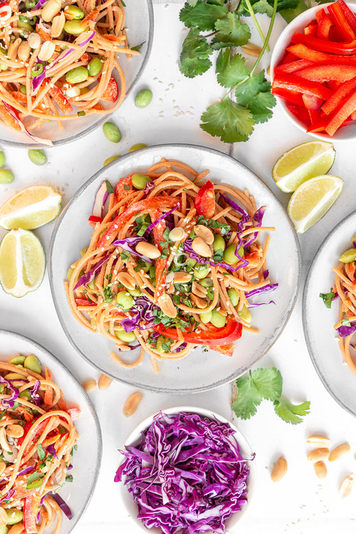 Colorful vegetable salad with noodles on a white plate, surrounded by ingredients like purple cabbage, red bell peppers, and lime wedges.