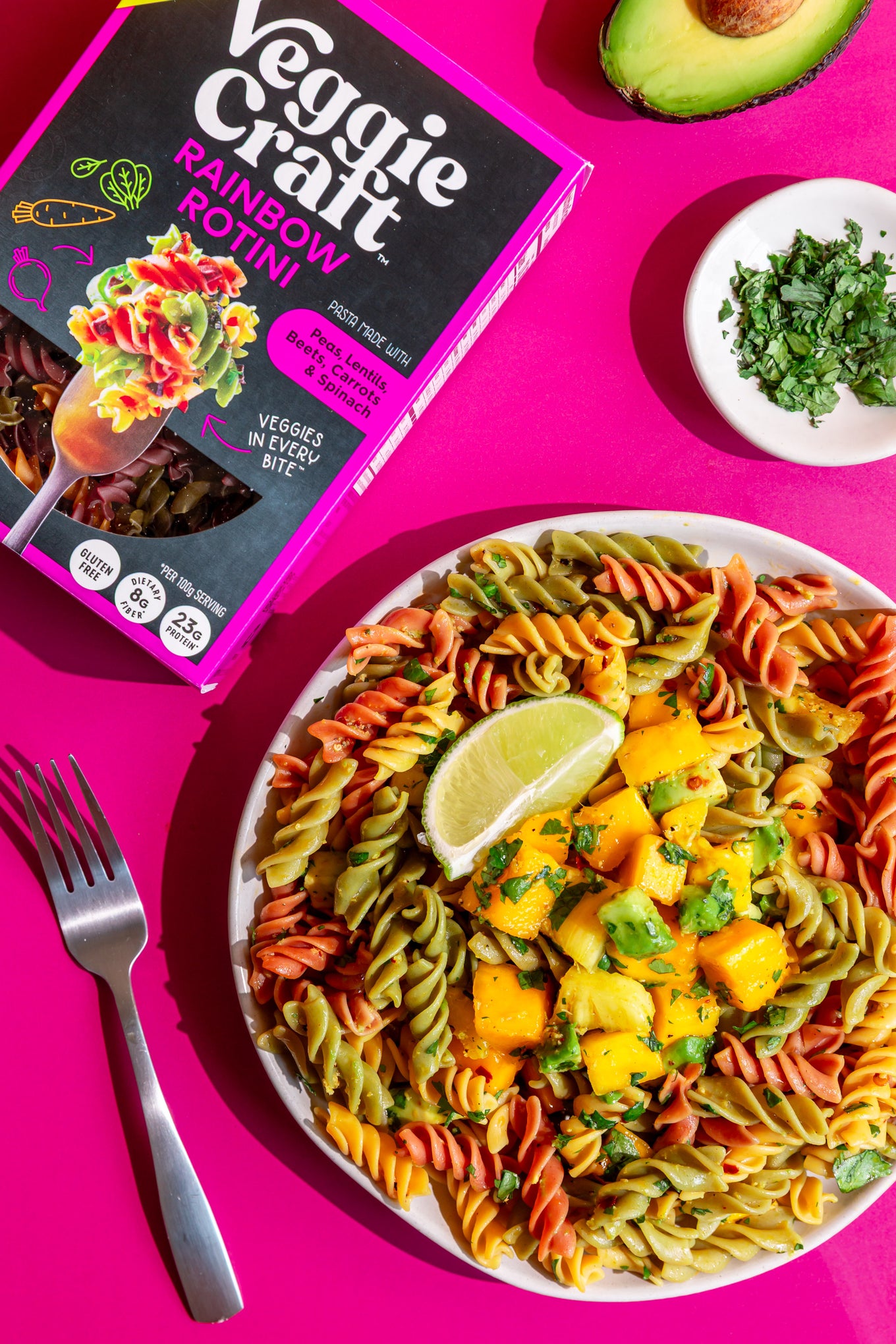 A plate of Veggie Craft Rainbow Rotini pasta with mango chunks, lime, and cilantro on a bright pink background, next to the product box and a fork.