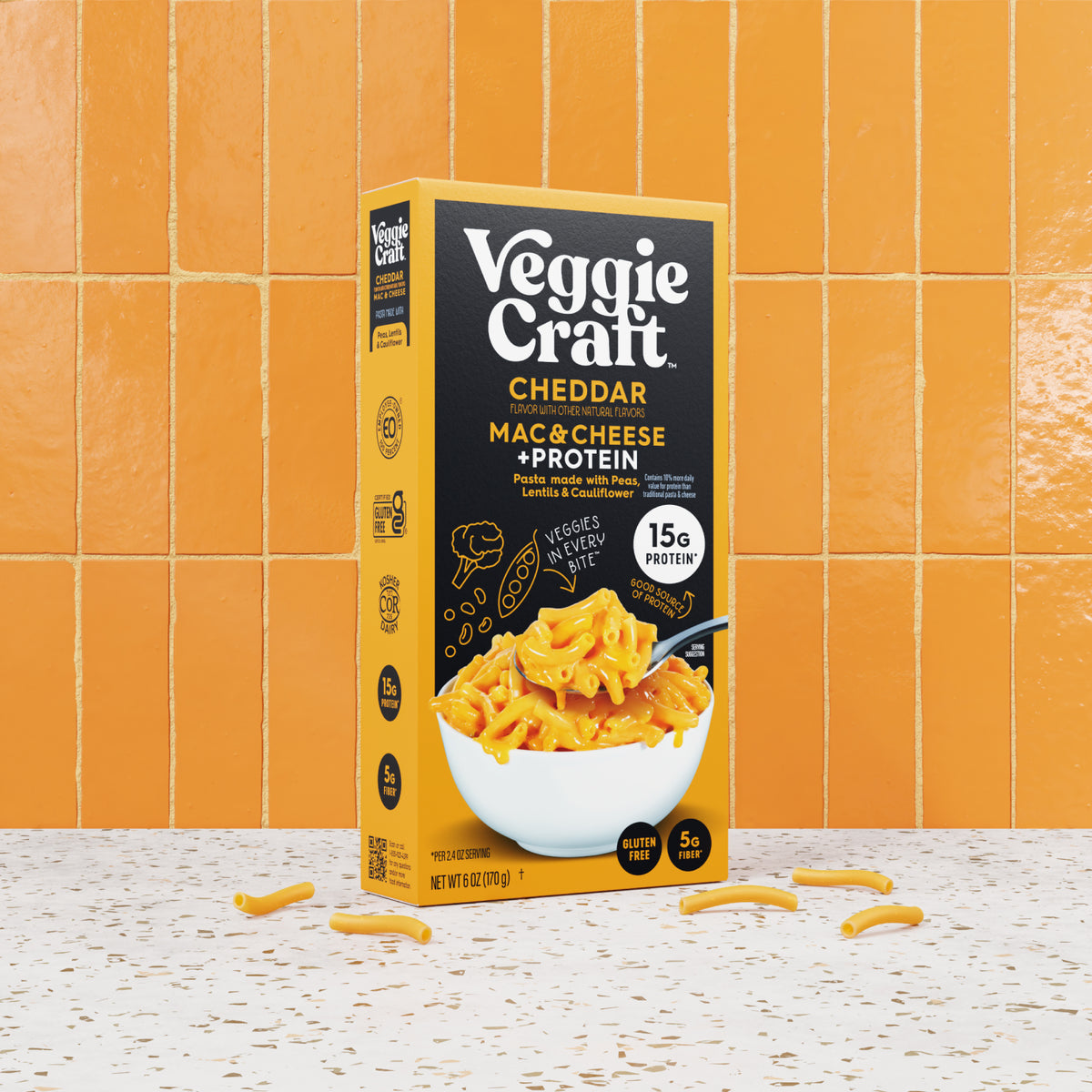 A box of Veggie Craft Cheddar Mac & Cheese + Protein stands on a speckled white surface with scattered cheddar curls in front, against a glossy orange tile background.