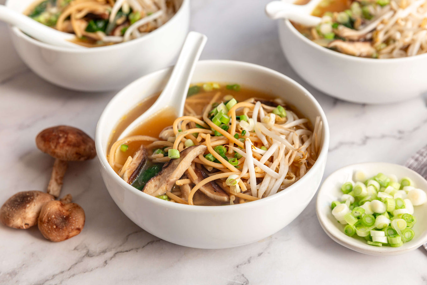 Bowl of noodle soup with a spoon, surrounded by mushrooms and green onions on a marble surface