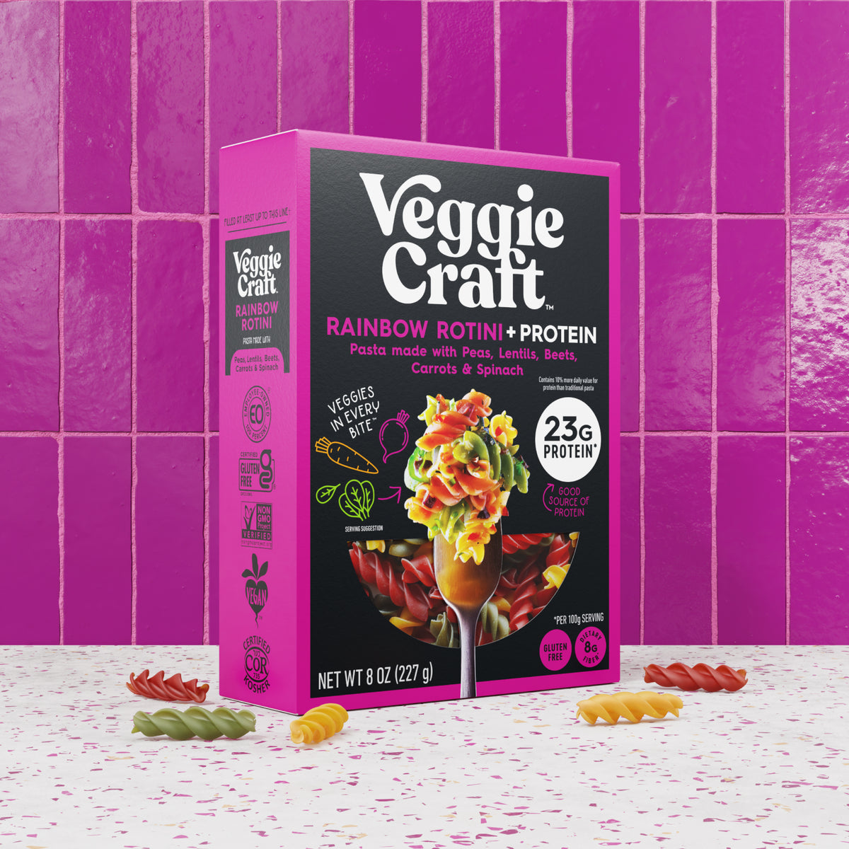 A box of Veggie Craft Rainbow Rotini + Protein pasta stands on a white speckled surface against a bright pink tiled background. The package is pink and black with white text and an illustration of a pasta-filled glass.