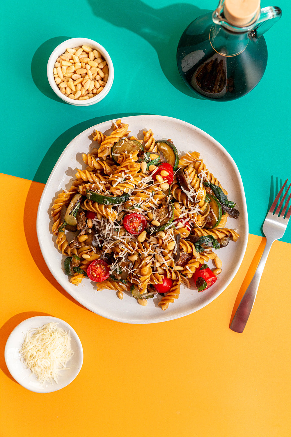 Pasta dish with vegetables on a white plate, surrounded by ingredients and a bottle of olive oil on a colorful background.