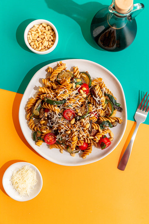 Pasta dish with vegetables on a white plate, surrounded by ingredients and a bottle of olive oil on a colorful background.