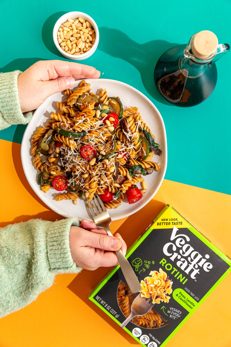 A plate of Veggie Craft Rotini pasta salad is held over a colorful split background, with a hand holding a fork and the Veggie Craft Rotini box visible in the foreground.