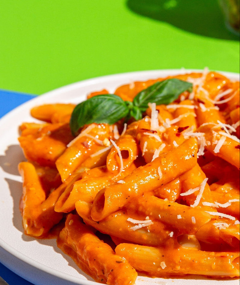 A white plate of penne pasta in a rich orange-red tomato sauce, garnished with fresh green basil leaves and shredded parmesan cheese, set against a vibrant blue and green background.
