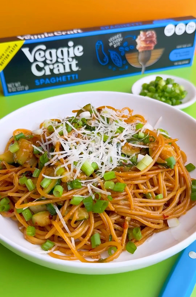 Spaghetti dish with green onions and parmesan cheese on a plate, with Veggie Craft spaghetti packaging in the background.